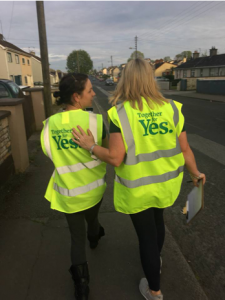 Two canvassers walk down a street with their backs to camera, wearing together for yes high vis vest. One has her arm on the other's back