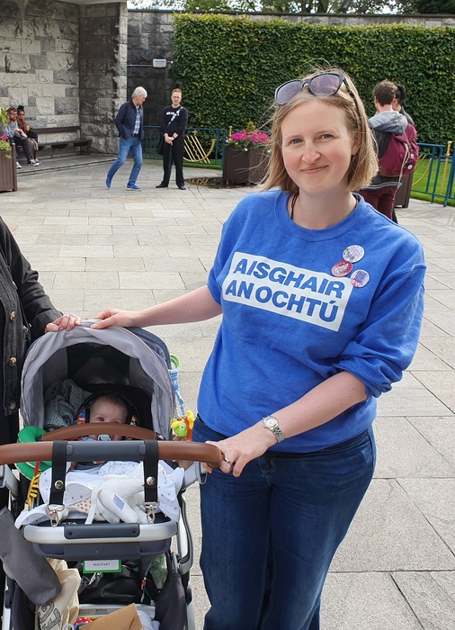 Photograph of Helen Stonehouse wearing a blue "Aisghair an Ochtú" sweater, pictured beside a baby in a buggy