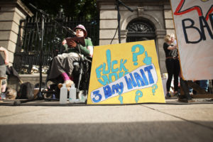 Image of a yellow placard that reads 'Fuck your 3 day wait'. In the background is Isolde ÓBrolcháin Carmody speaking at the 10th March for Choice last weekend.