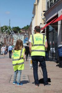 Sam Boland and his daughter, Ivy, standing with their backs to the camera, both wearing Together for Yes hi-vis vests.