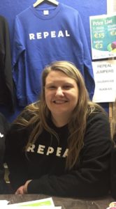 Darina Murray, ARC co-convener, is wearing a black REPEAL jumper and sitting at a table. There is a blue REPEAL jumper hanging up behind her. 
