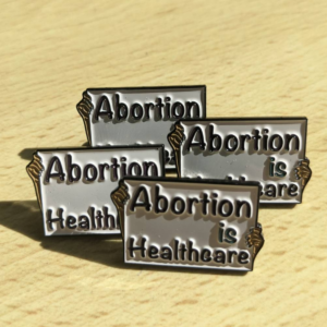 4 enamel pins against a pale wooden background. The pins are shaped like placards with a hand on either side holding it. It says "Abortion is Healthcare" against a white background.