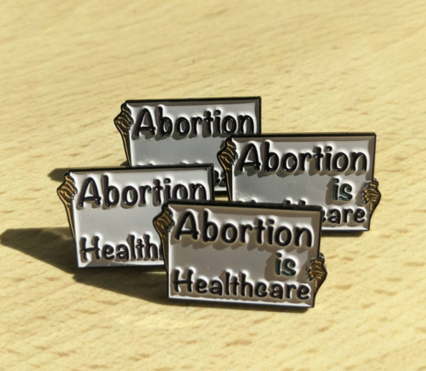 4 enamel pins against a pale wooden background. The pins are shaped like placards with a hand on either side holding it. It says "Abortion is Healthcare" against a white background.